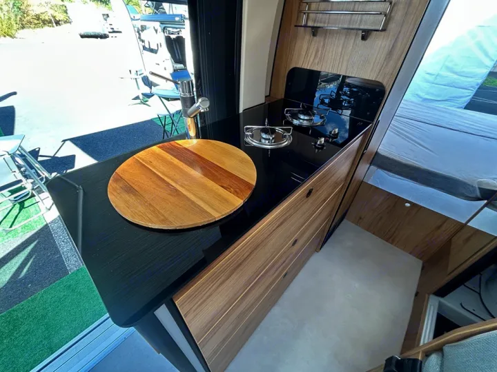 Modern rv rental kitchen with sleek black counter, two-burner stove, sink, wooden board, and warm wood cabinetry.. Font Vendôme Leader Camper Duo 2024