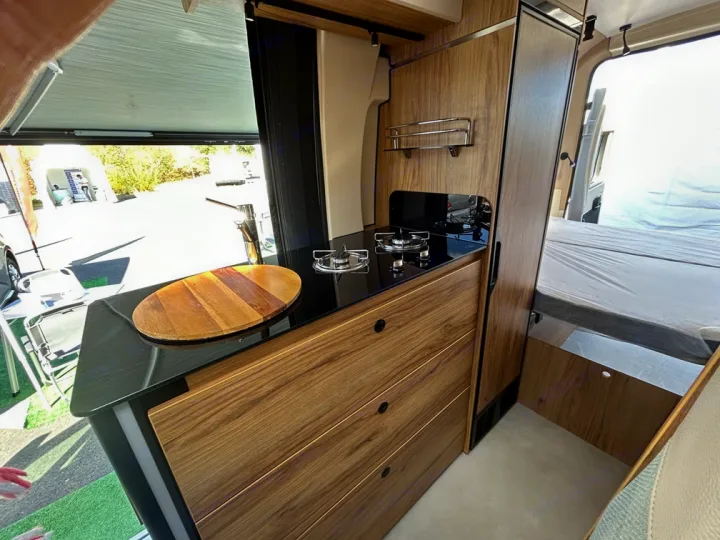 Modern rv rental interior: compact camper van kitchen with stovetop, sink, and wood cabinets. Bed and outdoor view visible.. Font Vendôme Leader Camper Duo 2024