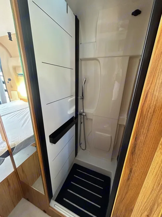 Compact shower stall in an RV rental, featuring a white interior, black mat, and reflections of the adjacent bedroom.. Font Vendôme Leader Camper Duo 2024