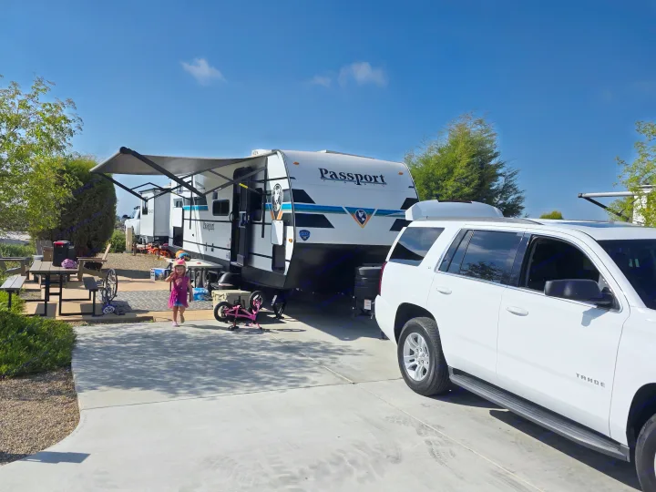 An RV rental adventure! A little girl in a pink helmet walks by a Passport travel trailer and white SUV at a sunny campsite.. Keystone RV Passport Classic 2025