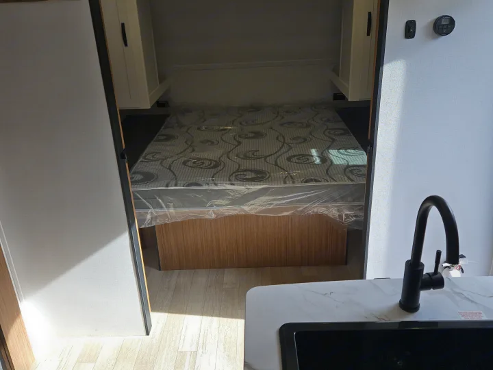 RV rental bedroom & kitchen view: new mattress with plastic, cabinets, a modern sink, and smart thermostat on light walls.. Keystone RV Passport Classic 2025