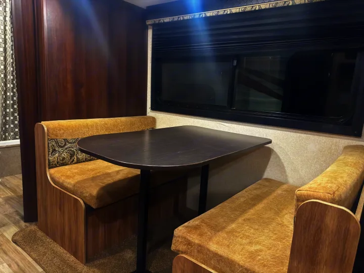 RV rental dinette interior: a dark table between two tan upholstered benches, with a window and horizontal blinds.. Jayco Jay Feather 2018