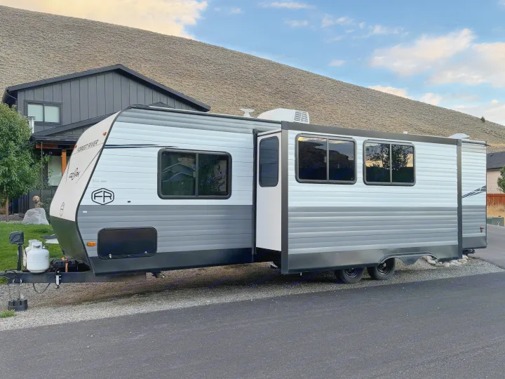 RV rental: A grey & white Forest River travel trailer with its slide-out extended, parked by a house & large hill.. Forest River 2690BHK Ozark 2025