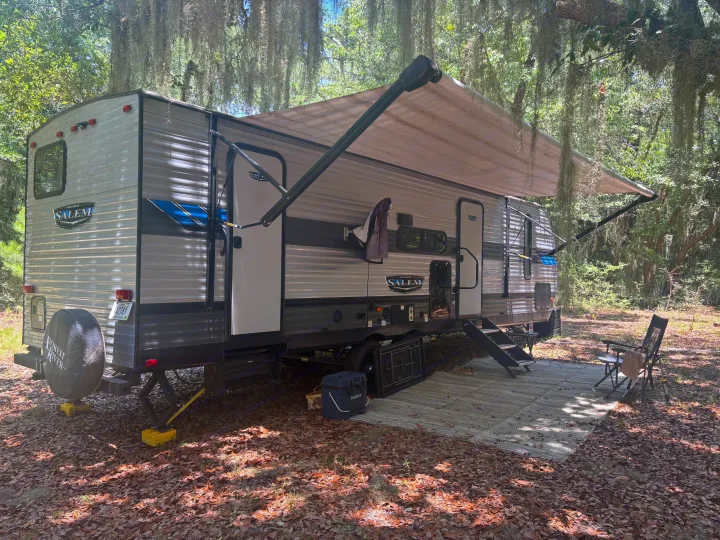 Forest River Salem rv rental travel trailer in a shaded, mossy campsite with an extended awning, outdoor mat, and chairs.. Forest River Salem 2022