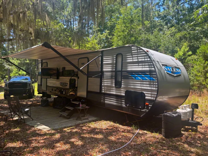 Travel trailer RV rental in a shaded forest campsite. An outdoor setup with a deployed awning, a GMC truck, and Spanish moss.. Forest River Salem 2022