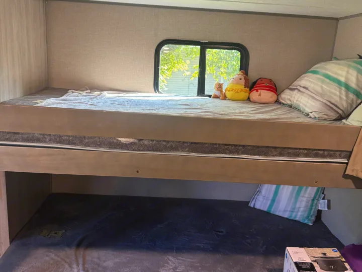 Inside an RV rental: cozy bunk beds. The top bunk has a window and plushies; the bottom has a blanket and slow cooker.. Coachmen Catalina summit series 8 2023