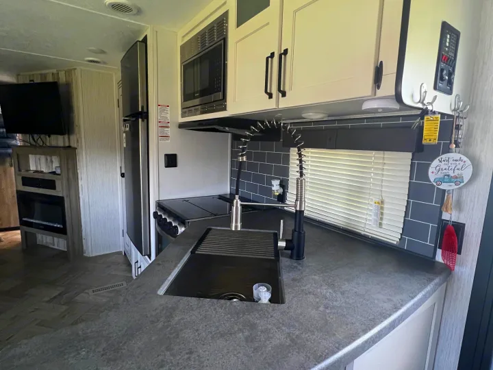 Modern RV rental kitchen with a black faucet, grey countertops, subway tile backsplash, microwave, fridge, and fireplace.. Forest River Palomino Puma 2021