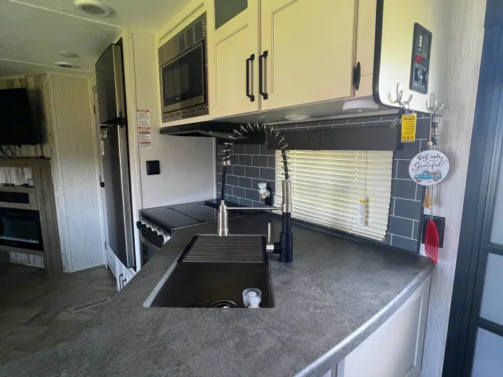 Modern RV rental kitchen featuring gray counters, dark sink, pull-down faucet, microwave, stove, and a fridge.. Forest River Palomino Puma 2021