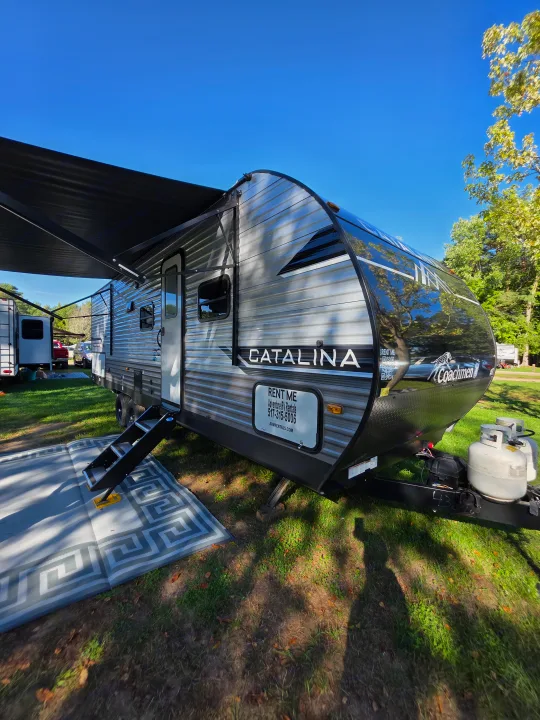 Sunny RV rental: a Catalina travel trailer with an extended awning and "Rent Me" sign, parked at a vibrant campsite.. Forest River Catalina 2024