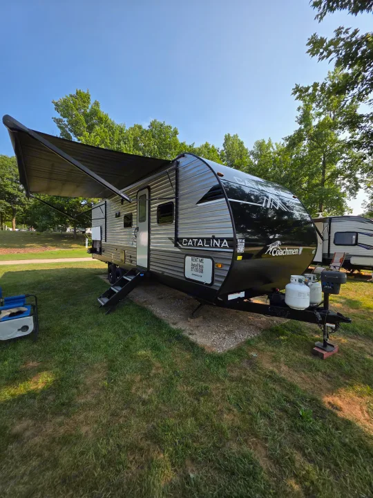 A Coachmen Catalina travel trailer for RV rental, parked at a sunny campsite with its awning extended under a clear blue sky.. Forest River Catalina 2024