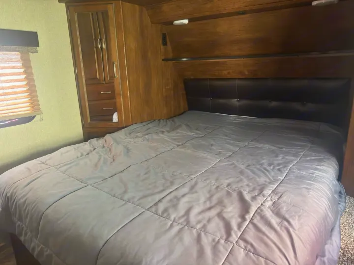 Comfortable RV rental bedroom featuring a large bed with a gray comforter, black headboard, and wood cabinetry.. Grand Design Reflection 2019