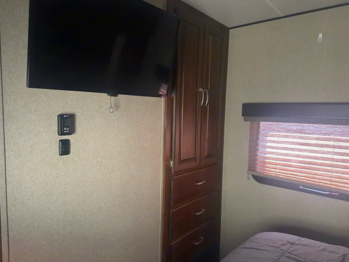 Interior of an RV rental bedroom showing a wall-mounted TV, dark wood cabinet, and a window with blinds over a bed.. Grand Design Reflection 2019