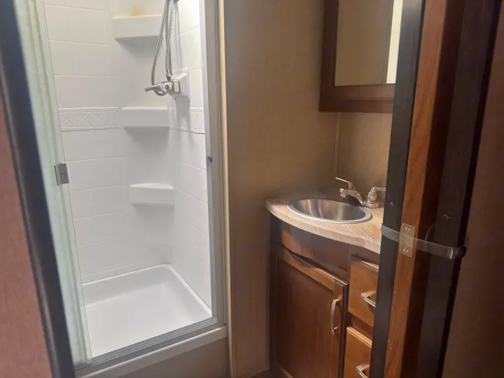Compact RV rental bathroom featuring a bright shower with glass door, vanity with a stainless steel sink, and mirror cabinet for storage.. Grand Design Reflection 2019