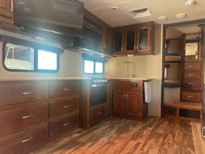 Spacious RV rental kitchen with modern wood cabinetry, oven, microwave, TV, and bunk beds visible through the doorway.. Grand Design Reflection 2019