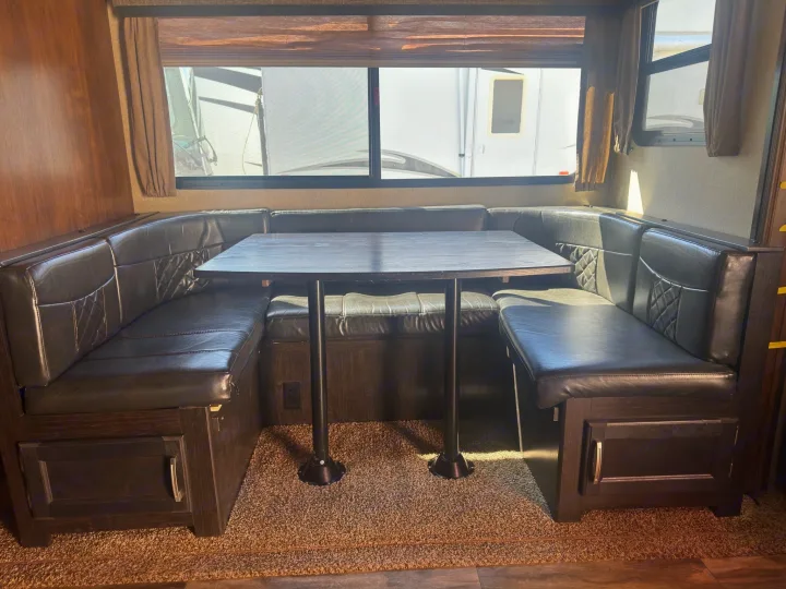 RV rental interior shows a spacious U-shaped dinette booth with dark cushions, a table, and convenient under-seat storage.. Grand Design Reflection 2019
