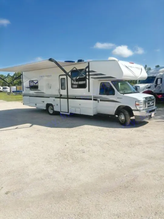 This white Class C RV rental, with its awning extended, is parked on a gravel lot under a clear blue sky. Ready for your trip!. Coachmen Freelander Freelander 2022