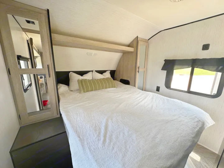 Inside an RV rental bedroom: a comfy bed with pillows, a window revealing nature, and wood-paneled storage.. Heartland RVs Prowler 2022