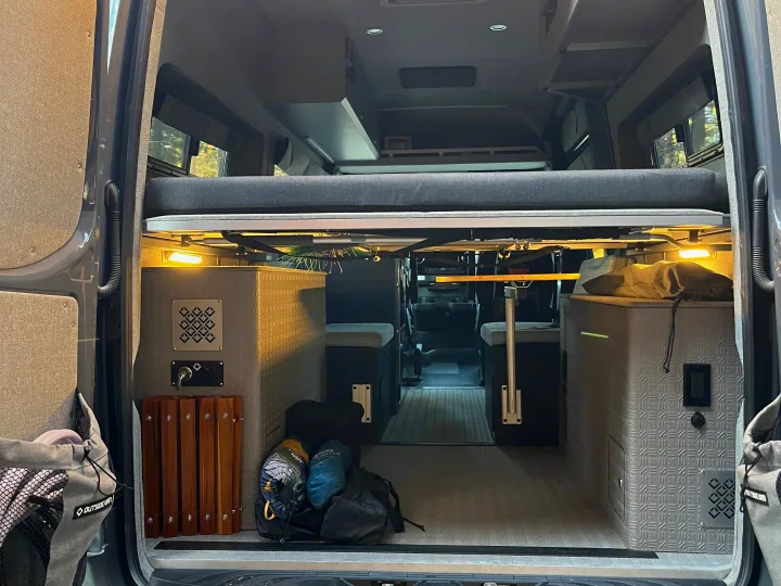 Rear view of an rv rental camper van, revealing a raised bed, under-bed storage, seating, and lighting for travel comfort.. Mercedes Benz Sprinter 144 High Roof 4WD 2020