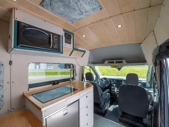 Inside a spacious RV rental: kitchen features microwave, sink, stovetop, fridge, and a view into the driver's cabin.. Ford Transit 2023