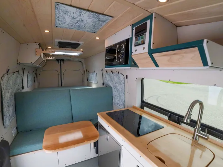 This RV rental interior features a compact kitchenette with sink & cooktop, overhead microwave, and a teal cushioned seating/bed area.. Ford Transit 2023