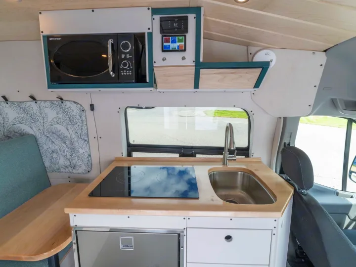 RV rental's compact kitchen and dining area: features microwave, induction cooktop, sink, fridge, light wood, and digital controls.. Ford Transit 2023