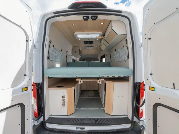 Open rear doors of a white RV rental reveal a custom camper van interior with a light blue bed, wood ceiling, and under-bed storage.. Ford Transit 2023