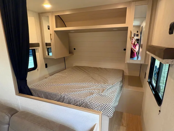 This RV rental features a comfortable bedroom with a checkered bed, built-in storage, and side windows.. Jayco Jay Flight 2027