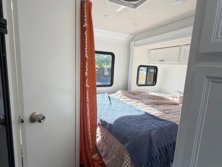 This inviting RV rental bedroom features a cozy bed with layered blankets, small windows, and a vibrant orange privacy curtain.. Thor Quantum 2018