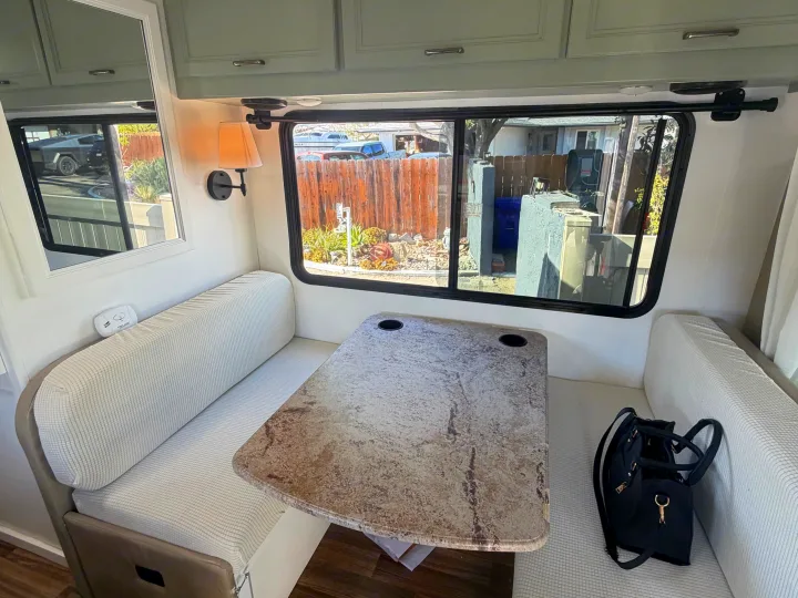 Cozy RV rental dinette featuring light-colored bench seating, a granite-look table, and an exterior view through the large window.. Thor Quantum 2018