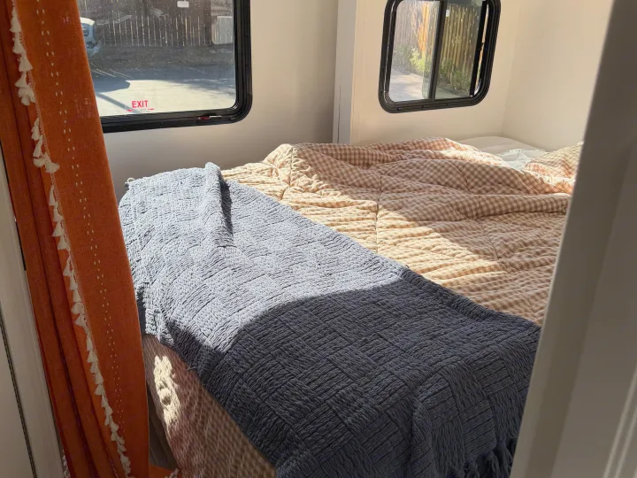 Cozy RV rental bedroom featuring a gingham comforter, textured grey blanket, and bright sunlit windows with an exit sign.. Thor Quantum 2018