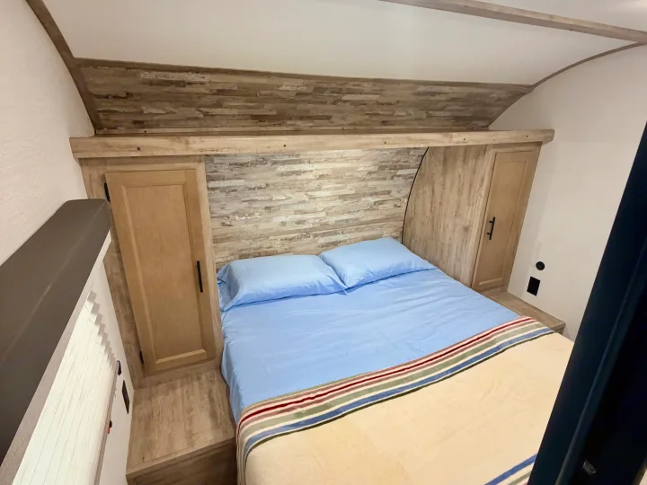 Inside a comfortable RV rental: a cozy bedroom features a bed with blue sheets, wood cabinets, and an accent wall.. Gulf Stream Enlighten SE 2025