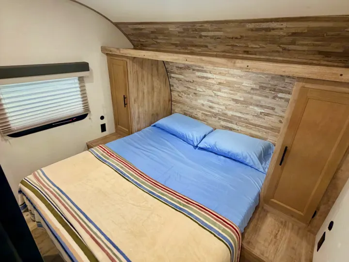 RV rental bedroom with a cozy bed, light blue sheets, striped blanket, and built-in wood-look storage.. Gulf Stream Enlighten SE 2025
