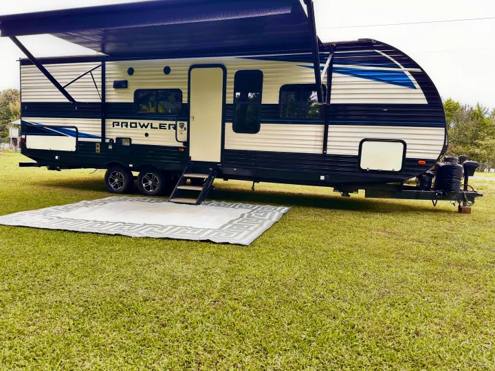 An RV rental, a Prowler travel trailer, set up for camping on green grass with its awning extended and a mat.. Heartland RVs Prowler 2022