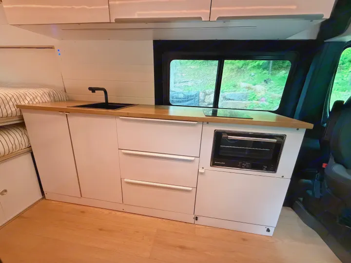 This rv rental's modern interior features a kitchenette with a sink, oven, and cooktop, beside a bed and window overlooking greenery.. RAM Promaster 2025