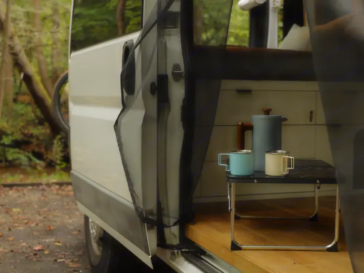 Morning coffee setup in an rv rental van: a grey pot, blue and cream mugs on a table. Nature outside.. Dodge Sprinter Van 2016