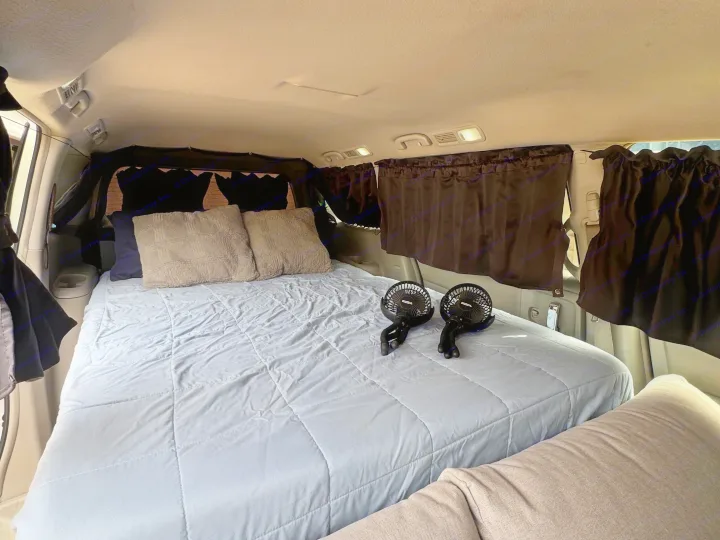 Inside a comfortable RV rental with a large bed, fluffy pillows, and personal fans, ideal for a road trip or van life.. Nissan Quest 2017