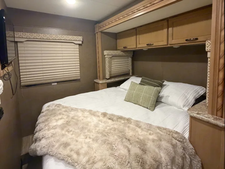 This RV rental bedroom offers a cozy bed with a furry throw, overhead cabinets, and window blinds for a relaxing stay.. Thor Freedom Elite 2017