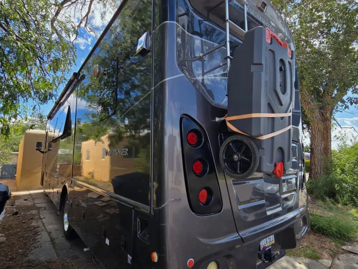 Side & rear view of a dark gray "Vacationer" RV rental, parked outdoors with a portable waste tank attached.. Holiday Rambler Vacationer 2019