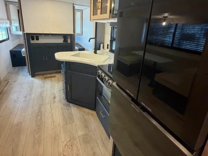 Modern RV rental kitchen with white sink, dark cabinets, stove, light wood-look flooring, and dinette reflection.. Avenger Avenger Trailer 2026