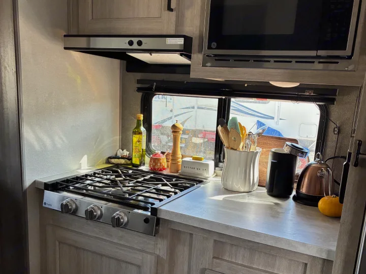 This bright RV rental kitchen features a gas stovetop, microwave, and a sunlit counter with cooking essentials like oil and spices.. Coachmen Catalina Summit 2024
