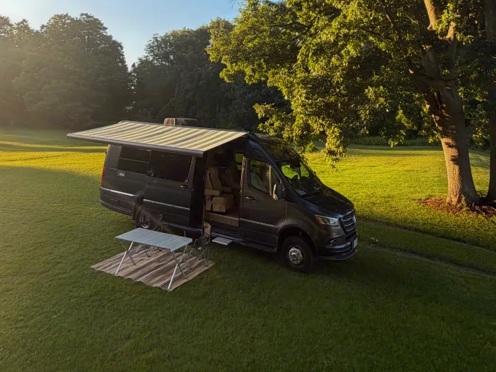 An RV rental Mercedes Sprinter camper van with its awning extended and camp setup, parked on lush grass.. Airstream Interstate 2025