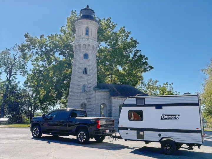 RV rental ready: Pickup truck with Coleman travel trailer parked at a grand stone lighthouse against a clear blue sky.. Keystone Coleman 13b 2027