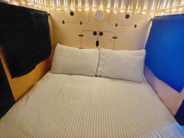 Cozy RV rental bed with striped bedding, string lights, and a unique bear-faced wooden panel overhead.. Hiker 5x8 Highway Deluxe 2025