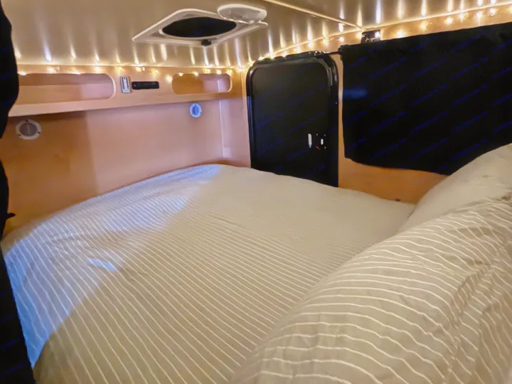 Cozy RV rental interior featuring a bed with striped sheets, ambient string lights, and built-in wall storage for a comfortable stay.. Hiker 5x8 Highway Deluxe 2025