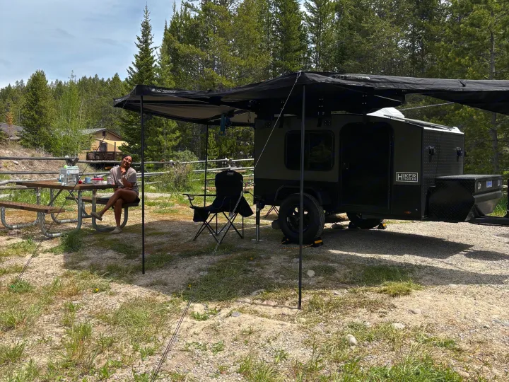 Enjoying nature with an **rv rental**! A woman relaxes by her Hiker Trailer camper, complete with an awning & picnic table, surrounded by pine trees.. Hiker 5x8 Highway Deluxe 2025