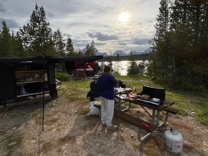Lakeside cooking during an RV rental trip, with mountain views and a cozy camper setup at a scenic campsite.. Hiker 5x8 Highway Deluxe 2025