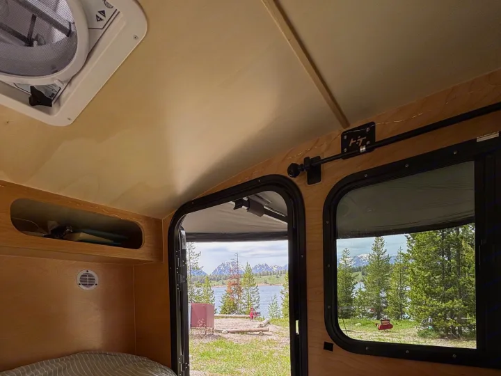 Inside a cozy RV rental: striped bed, open door, and window revealing a serene lake, lush trees, and majestic mountains.. Hiker 5x8 Highway Deluxe 2025