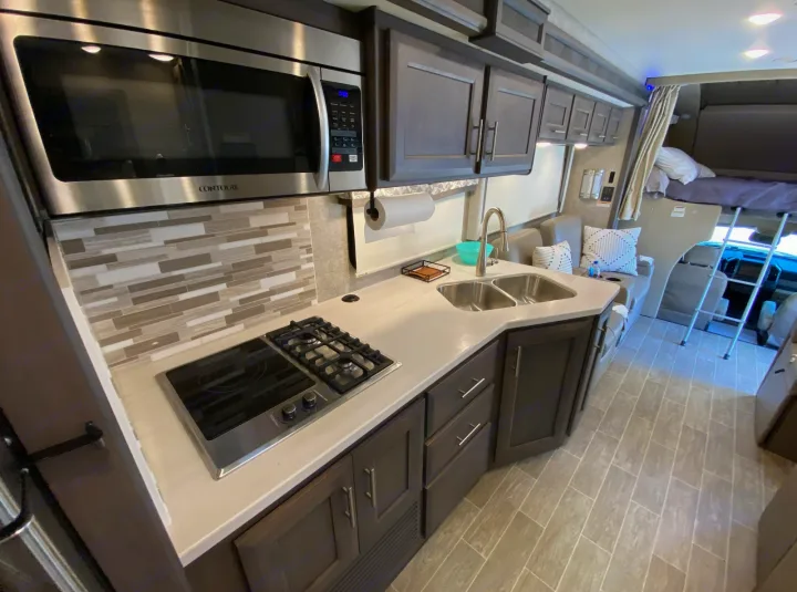 Modern rv rental kitchen with microwave, stove, sink, and sleek backsplash. Features cozy seating & over-cab bed area.. Thor Super C 2021