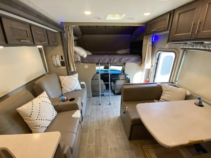 View inside an RV rental: living space with sofa, dinette with table, overhead bunk bed, and front cab area.. Thor Super C 2021