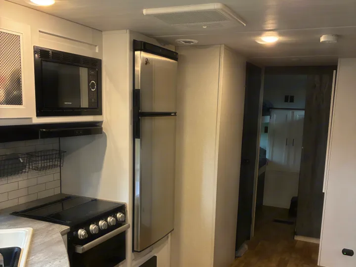 Modern RV rental kitchen with a stainless steel fridge, microwave, and stovetop. A hallway leads to a sleeping area. Clean design.. Keystone RV Hideout 2022